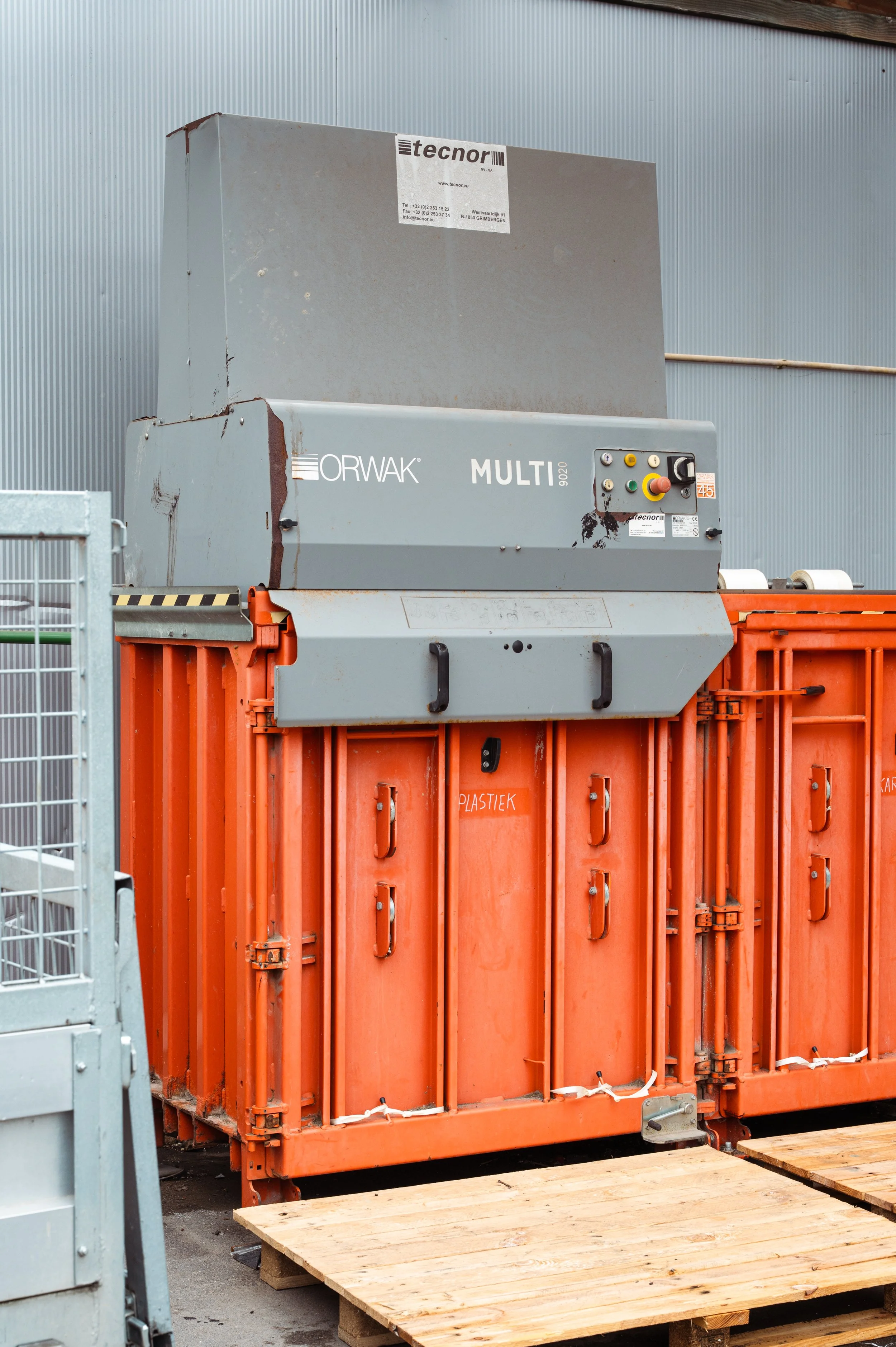 Een industriële afvalpers met een grote grijzer aluminium motor en een oranje afvalcontainer eronder, op houten pallets tegen een metalen wand.