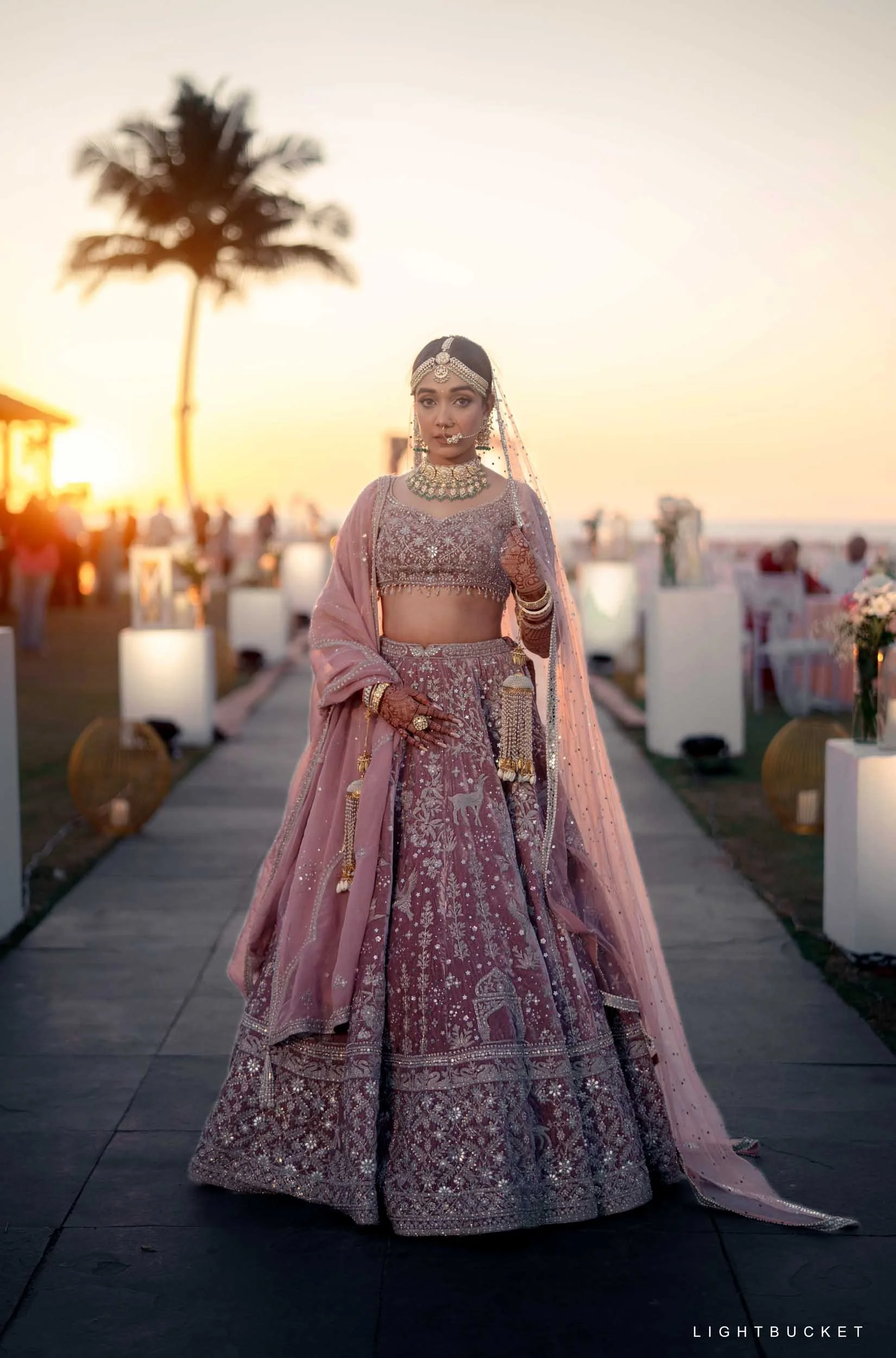 Woman in embellished Indian bridal attire with pink lehenga and jewelry, standing outdoors at sunset with palm tree in background.