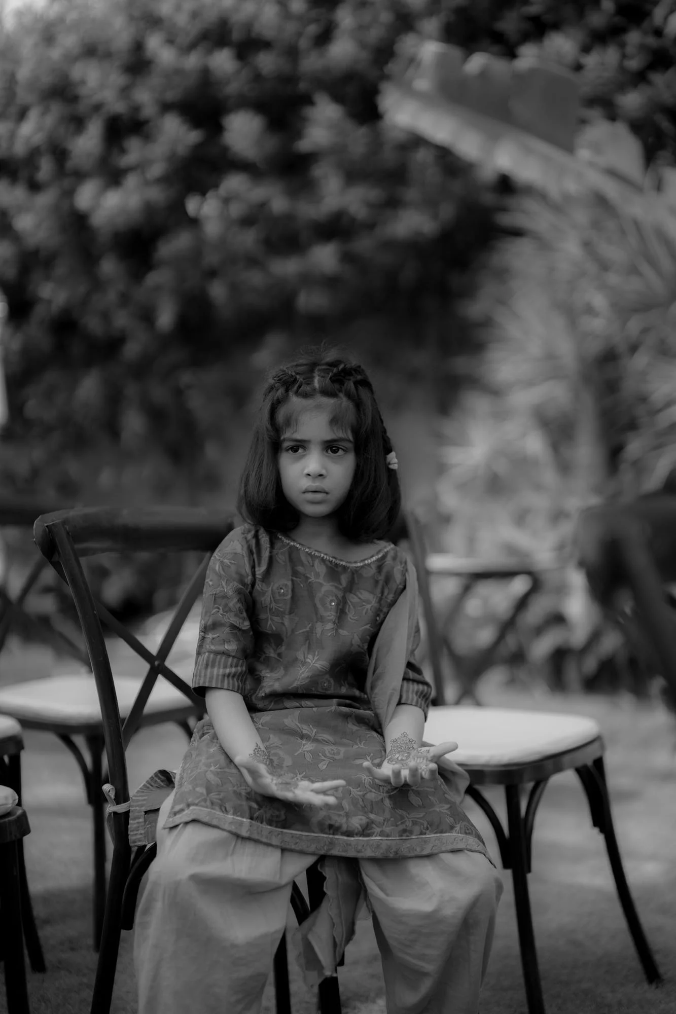 A young girl sitting on a chair outdoors with a serious expression, holding her hands with henna designs, surrounded by other chairs and greenery.