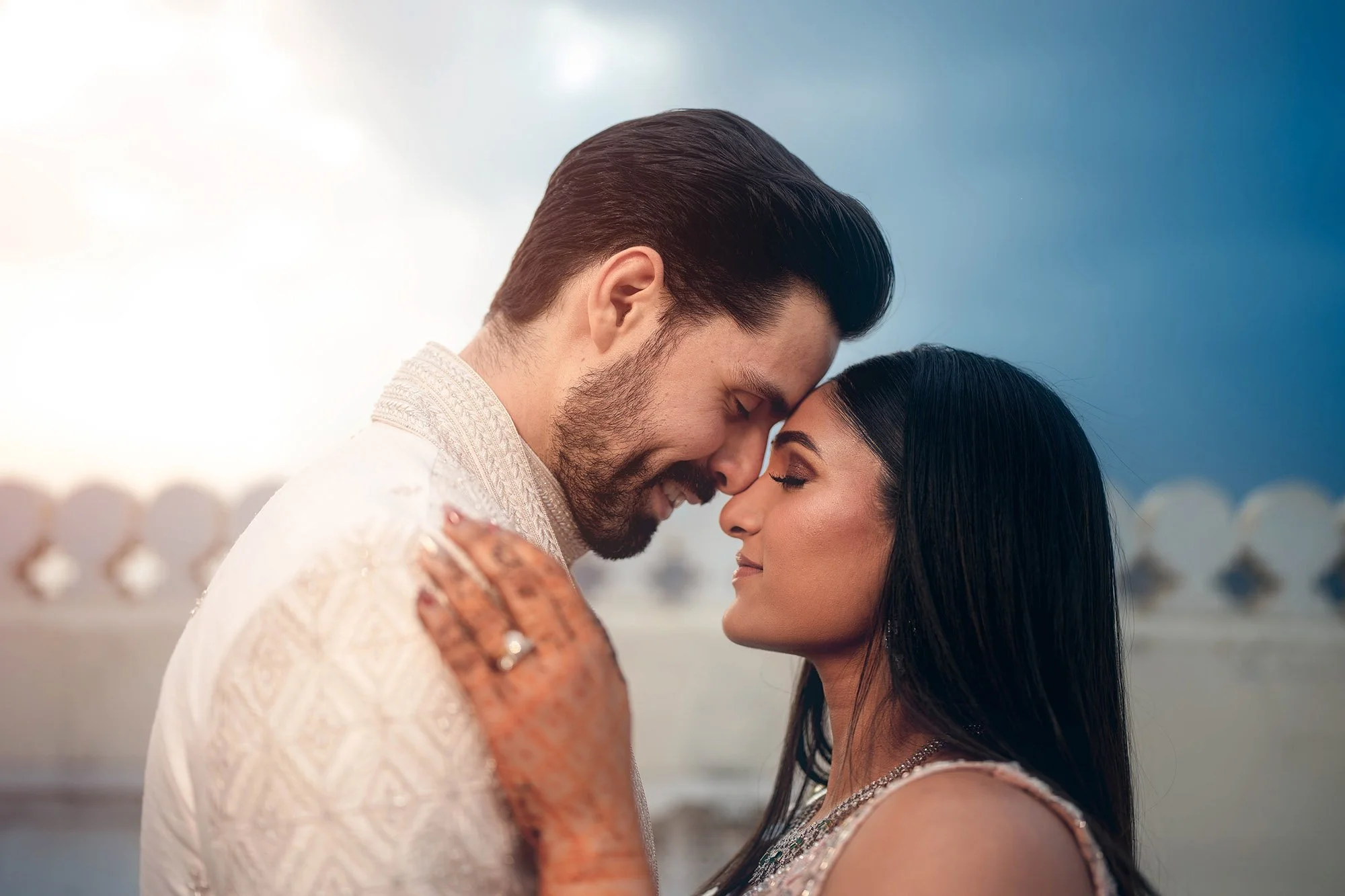 Couple embracing with foreheads touching, woman with henna on hand, wearing light-colored traditional attire, outdoors with sunlight in the background.