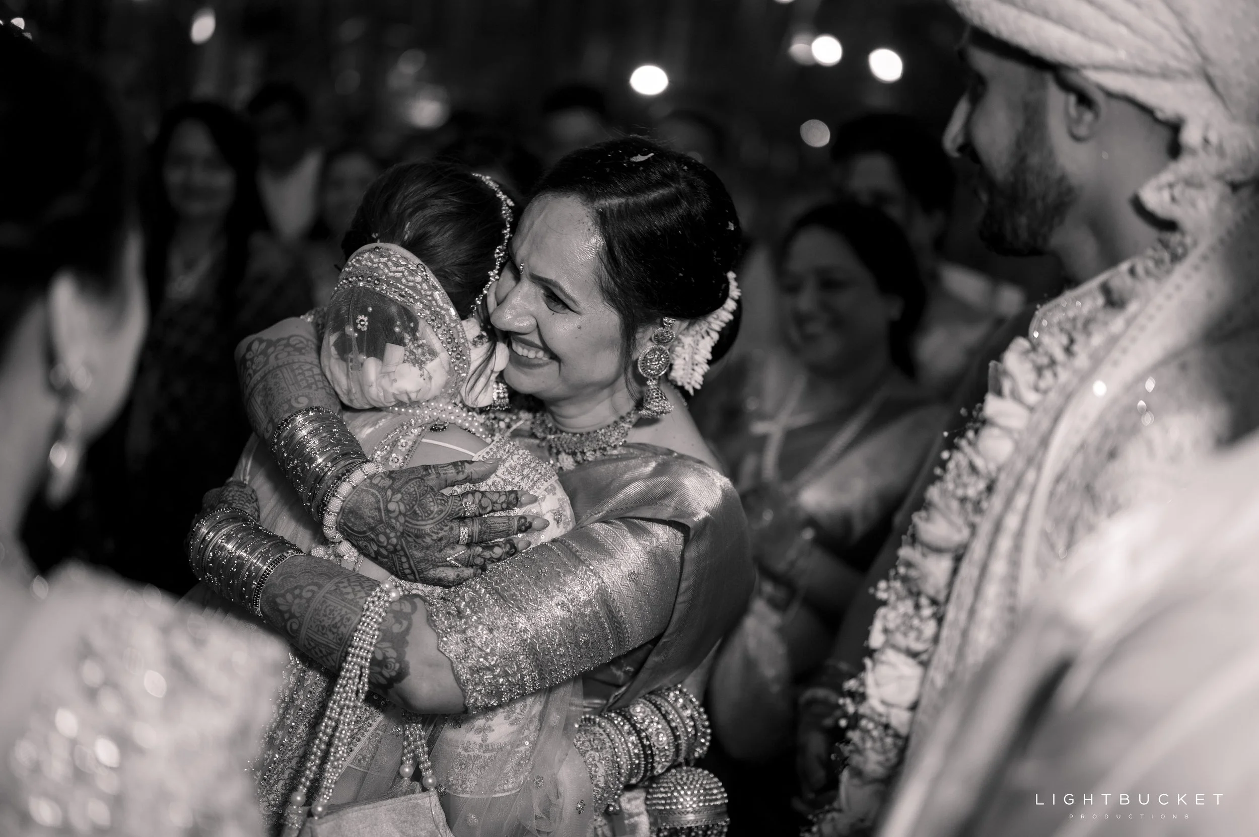A bride and a guest embracing at a traditional Indian wedding. The bride is smiling, dressed in intricate traditional attire with jewelry, while others in the background are also wearing traditional outfits, all in black and white.