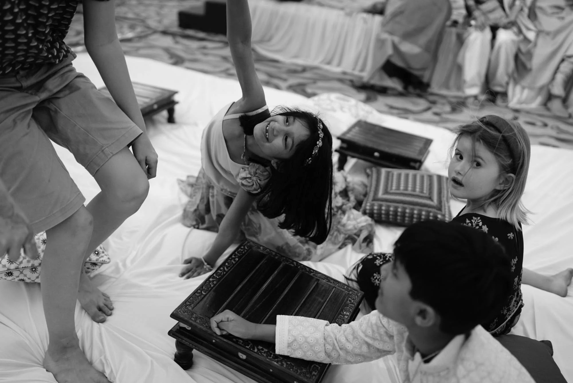 Group of children playing a board game on a bed, with some sitting and one girl kneeling and leaning over, with pillows and blankets around.