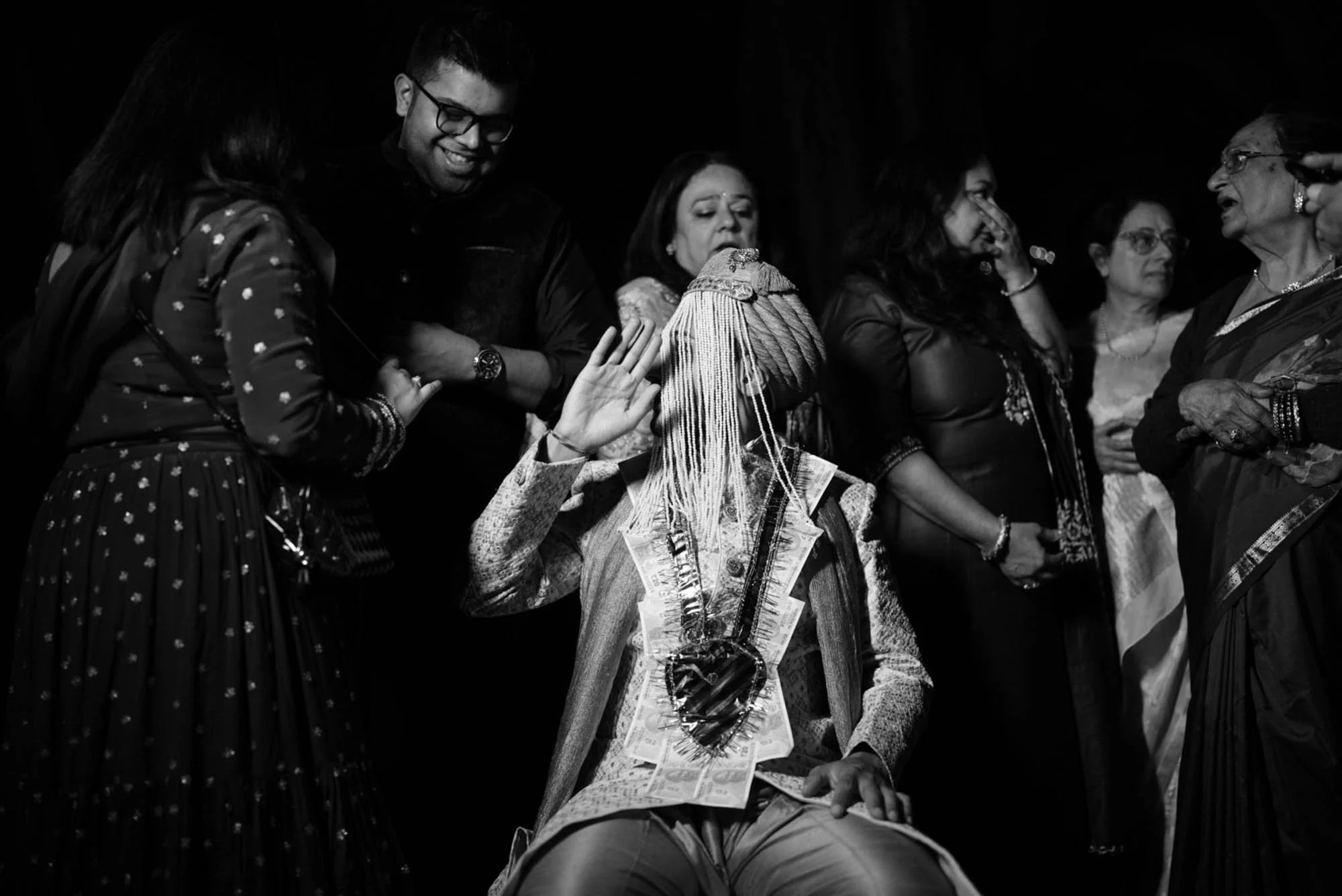Woman sitting with money and jewelry around her neck, surrounded by standing women, some with hands near face, on stage in a black and white photo.