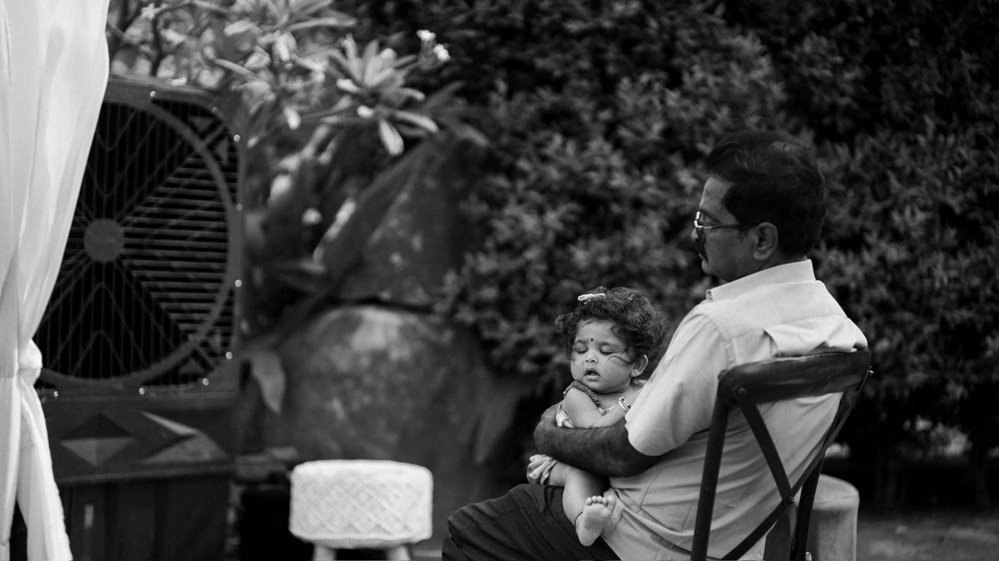 A man holding a sleepy young girl on his lap, sitting outdoors in a garden area with plants and a large decorative pot in the background.