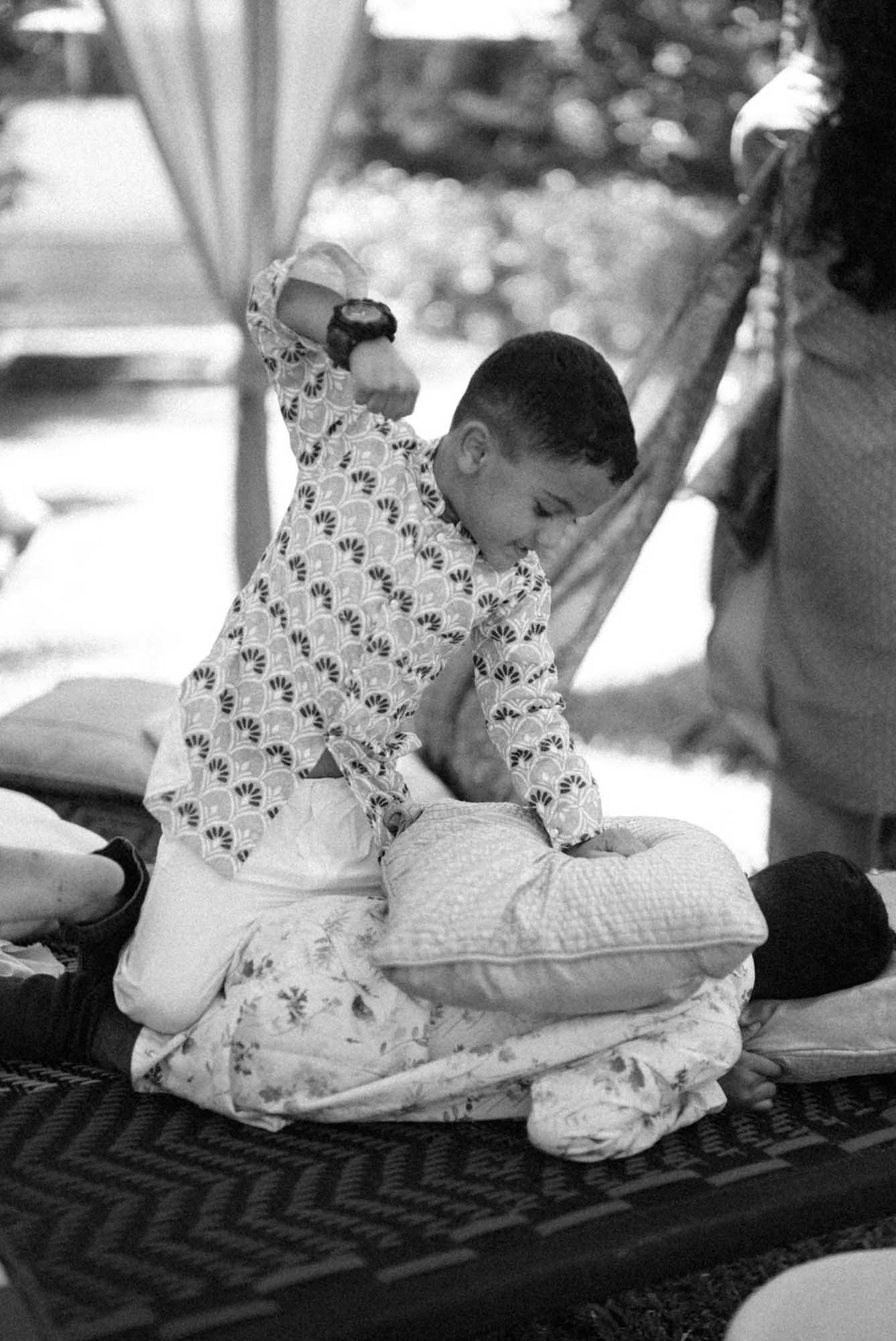 A young boy kneeling on a cushion, smiling, with his hand raised, in an outdoor setting during daytime.