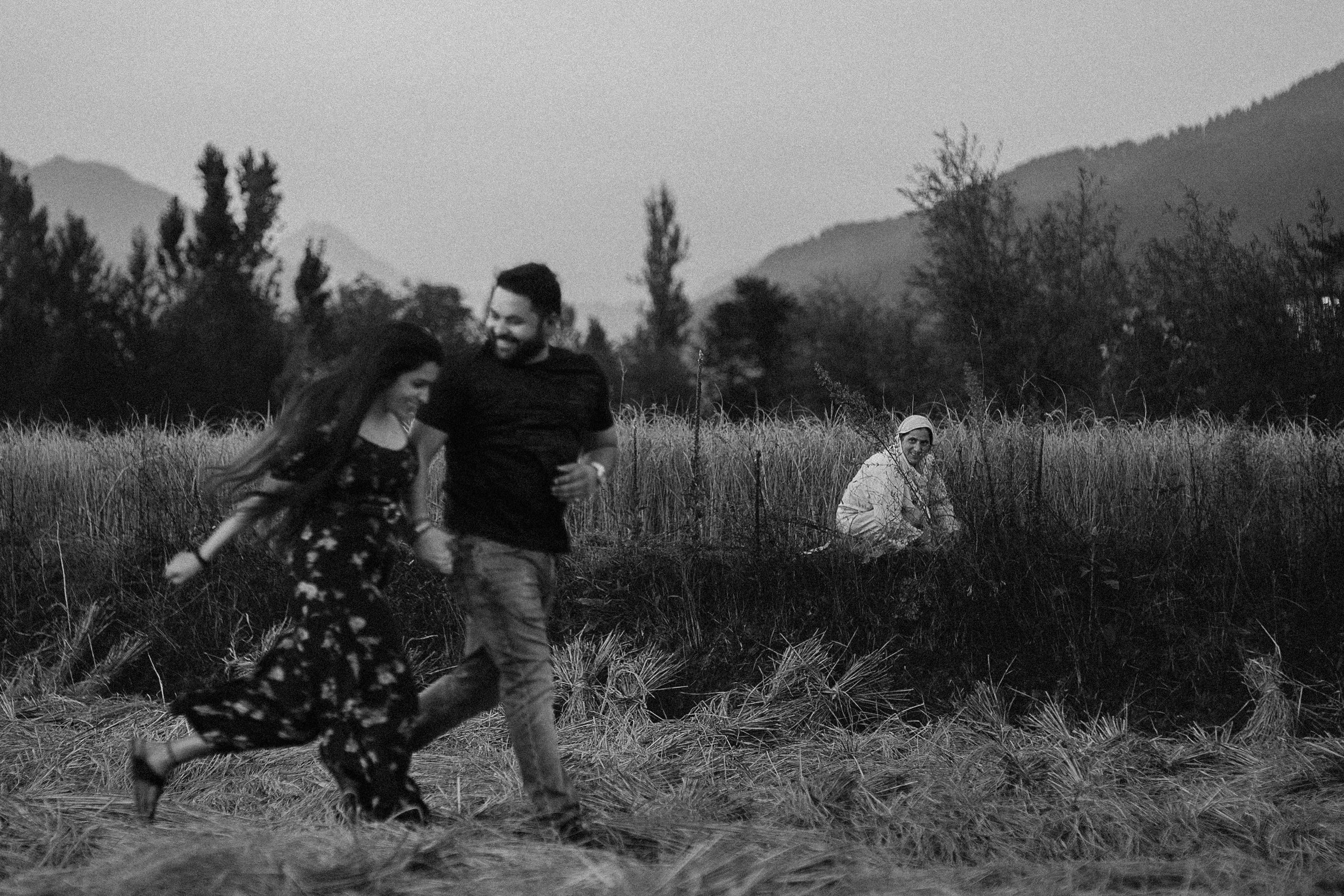 A man and woman running together through a field, while a woman sits nearby in the grass watching them.