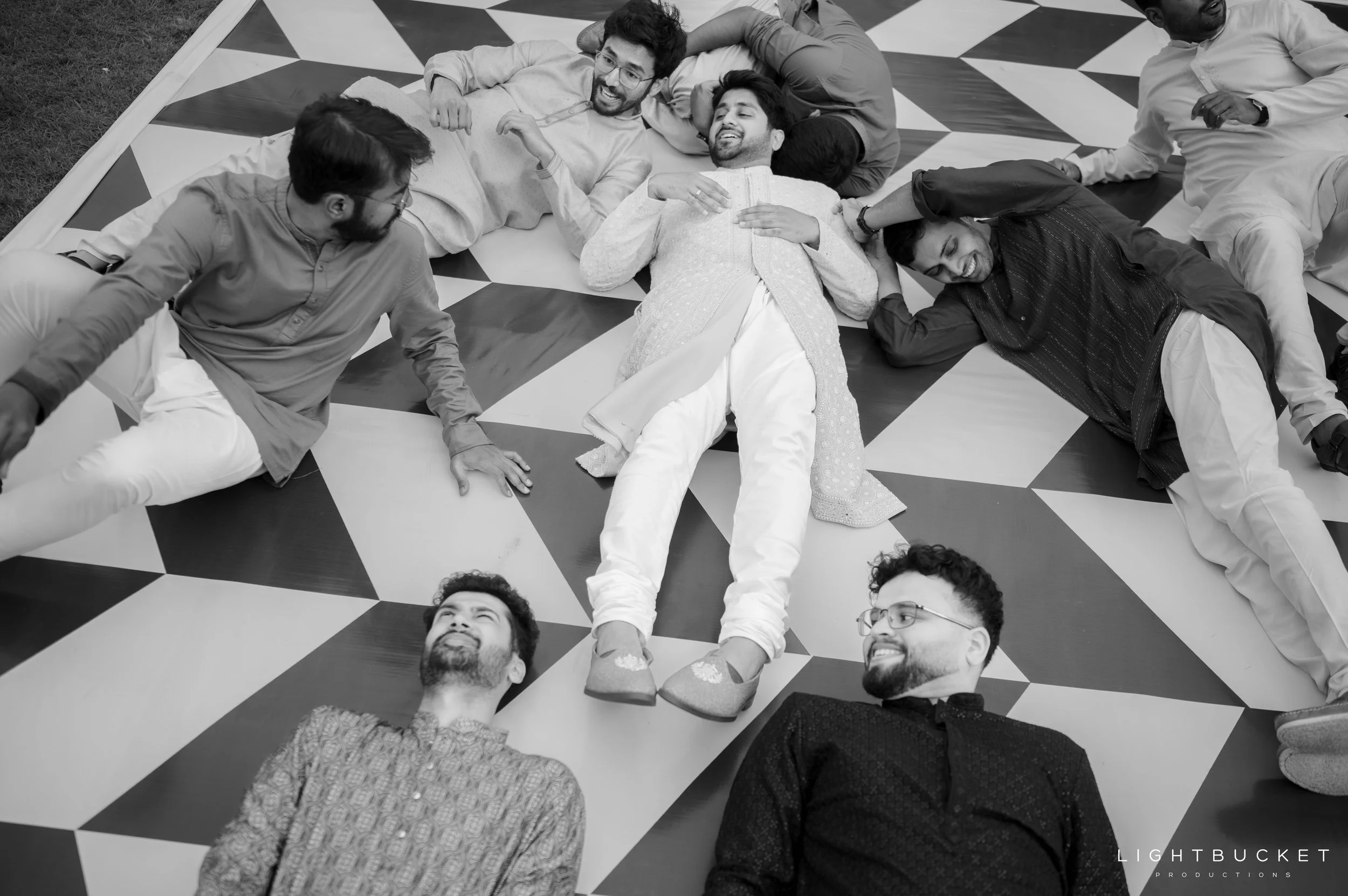 A group of nine people lying on a patterned floor, smiling and relaxing, with one person in the center wearing traditional Indian attire.