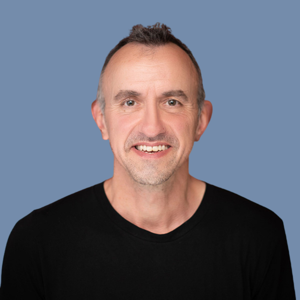 Headshot of smiling middle-aged man with short, slightly spiked brown hair, wearing a black shirt, set against a plain blue background.