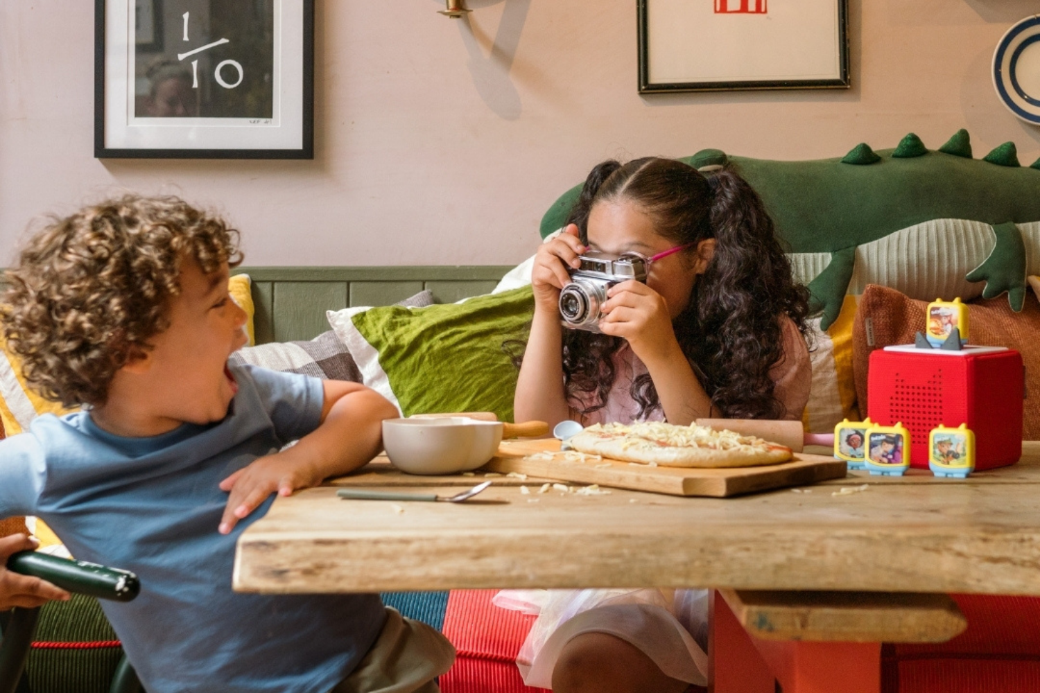 A boy with curly hair laughing with a girl taking a photo with a camera at a table with a pizza, bowls, and utensils, in a room with colorful decorations and framed pictures on the wall with audio drama in the background.