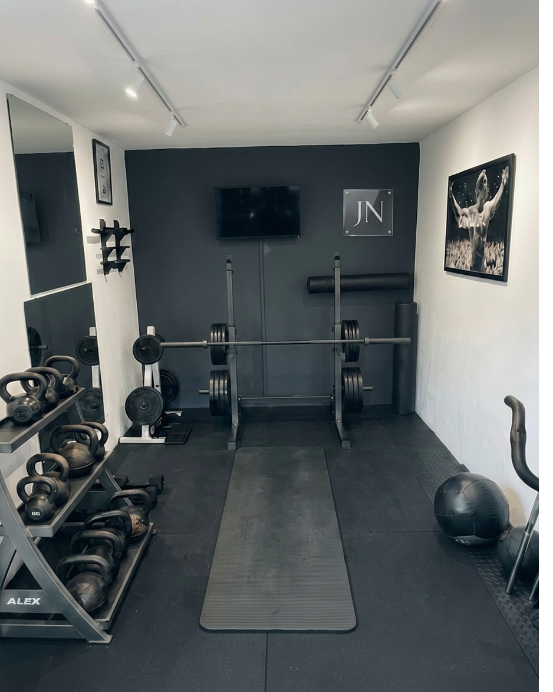 Home gym with black weight plates on a barbell, kettlebells, dumbbells, a medicine ball, and exercise mats, with a TV and motivational posters on the wall.