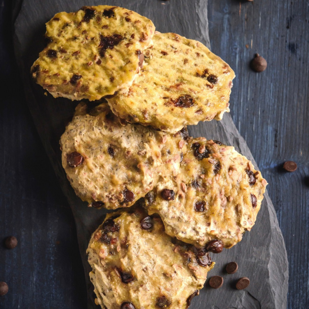 Four chocolate chip cookies on a black slate serving board on a dark wooden surface.