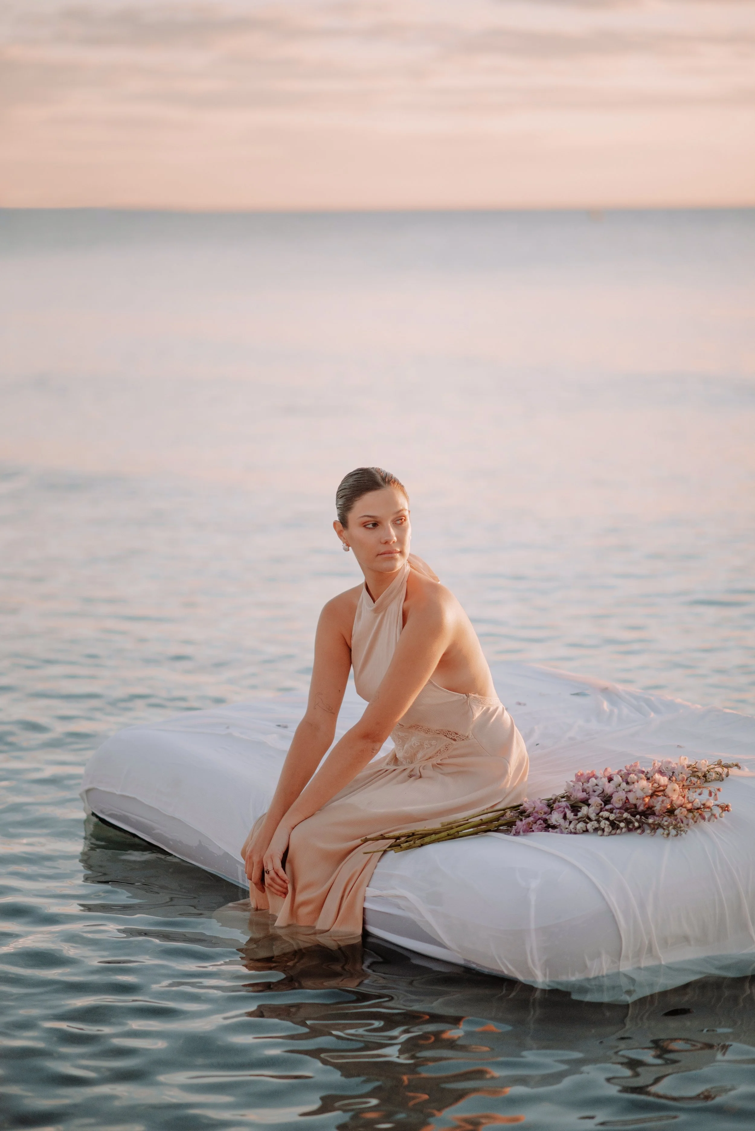 Mujer sentada en cama flotante en el agua con un ramo de flores, durante el atardecer en el mar.