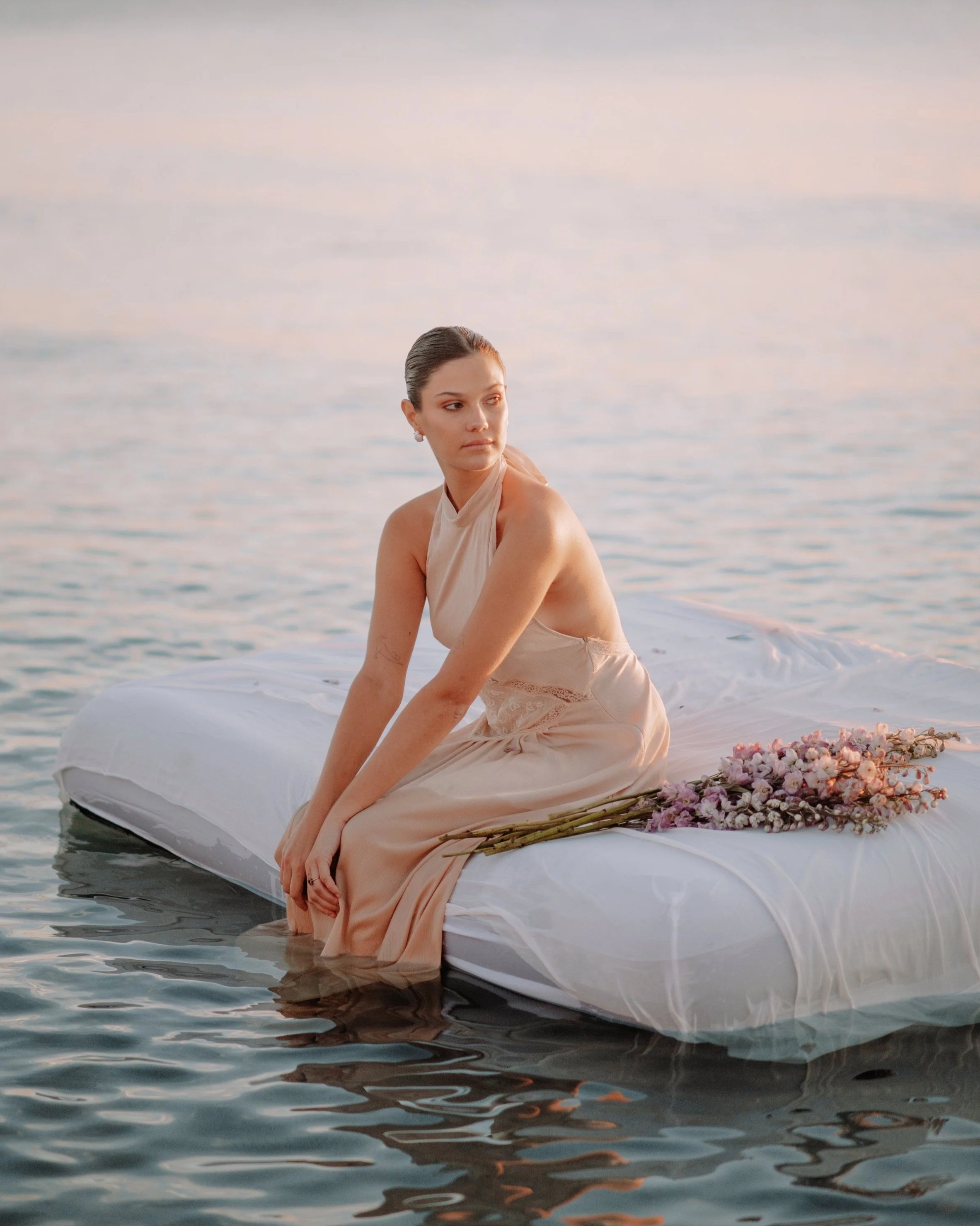 Mujer con vestido sentado en una cama flotante en el agua con un ramo de flores a su lado durante el atardecer.