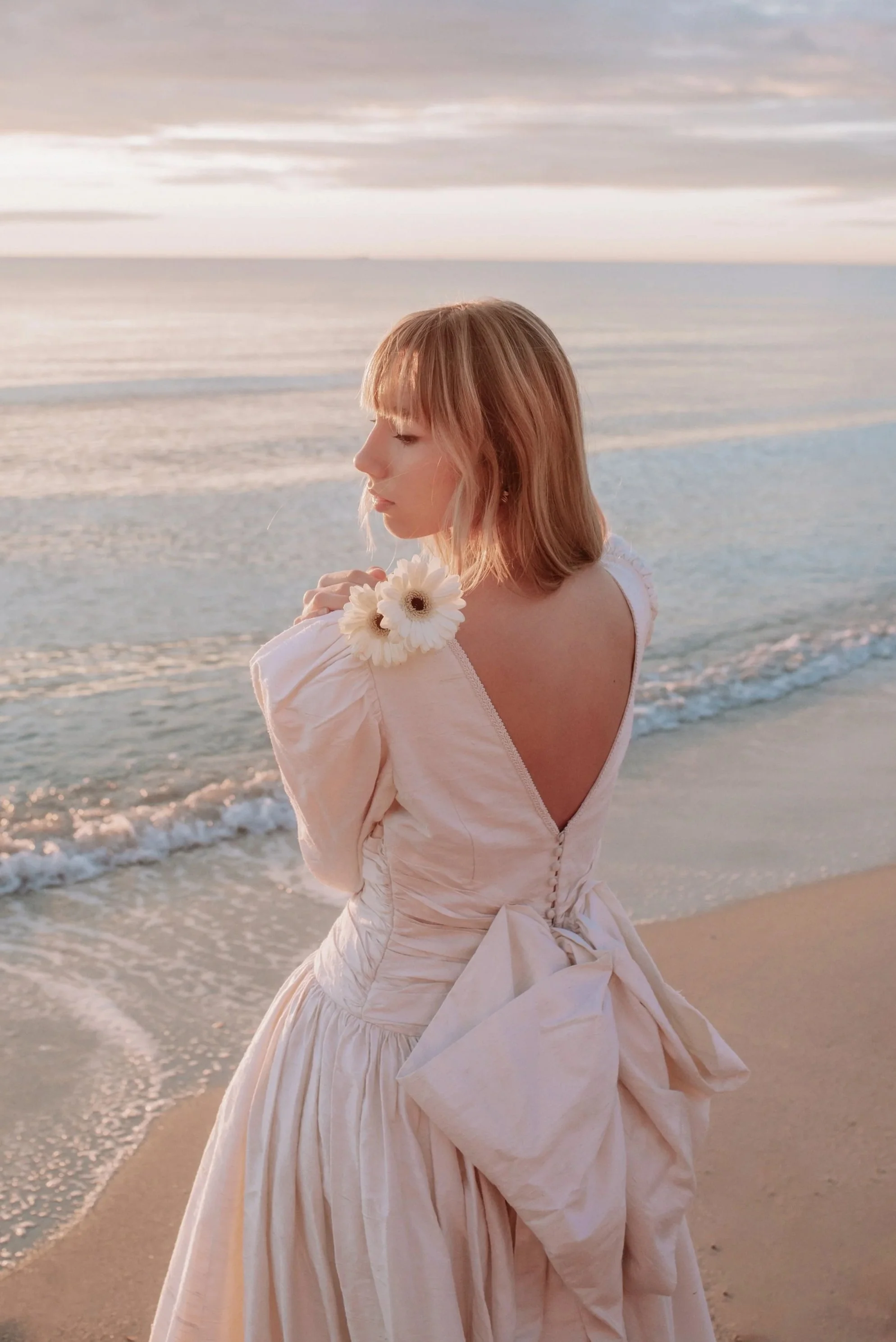 Una mujer con vestido largo y flor en el hombro, de cabello rubio y corto, de perfil, en la playa durante el atardecer