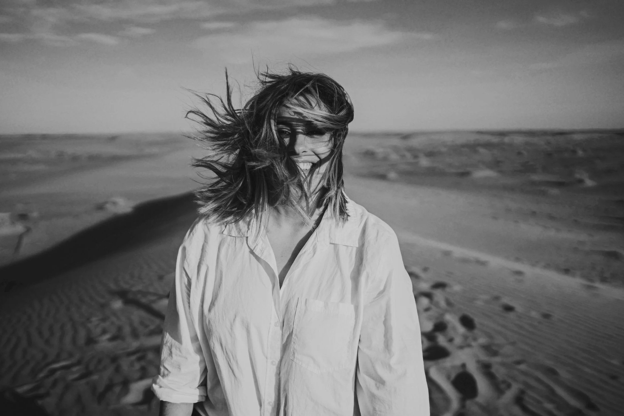 Mujer sonriendo en una playa arenosa con viento en el cabello, en blanco y negro.