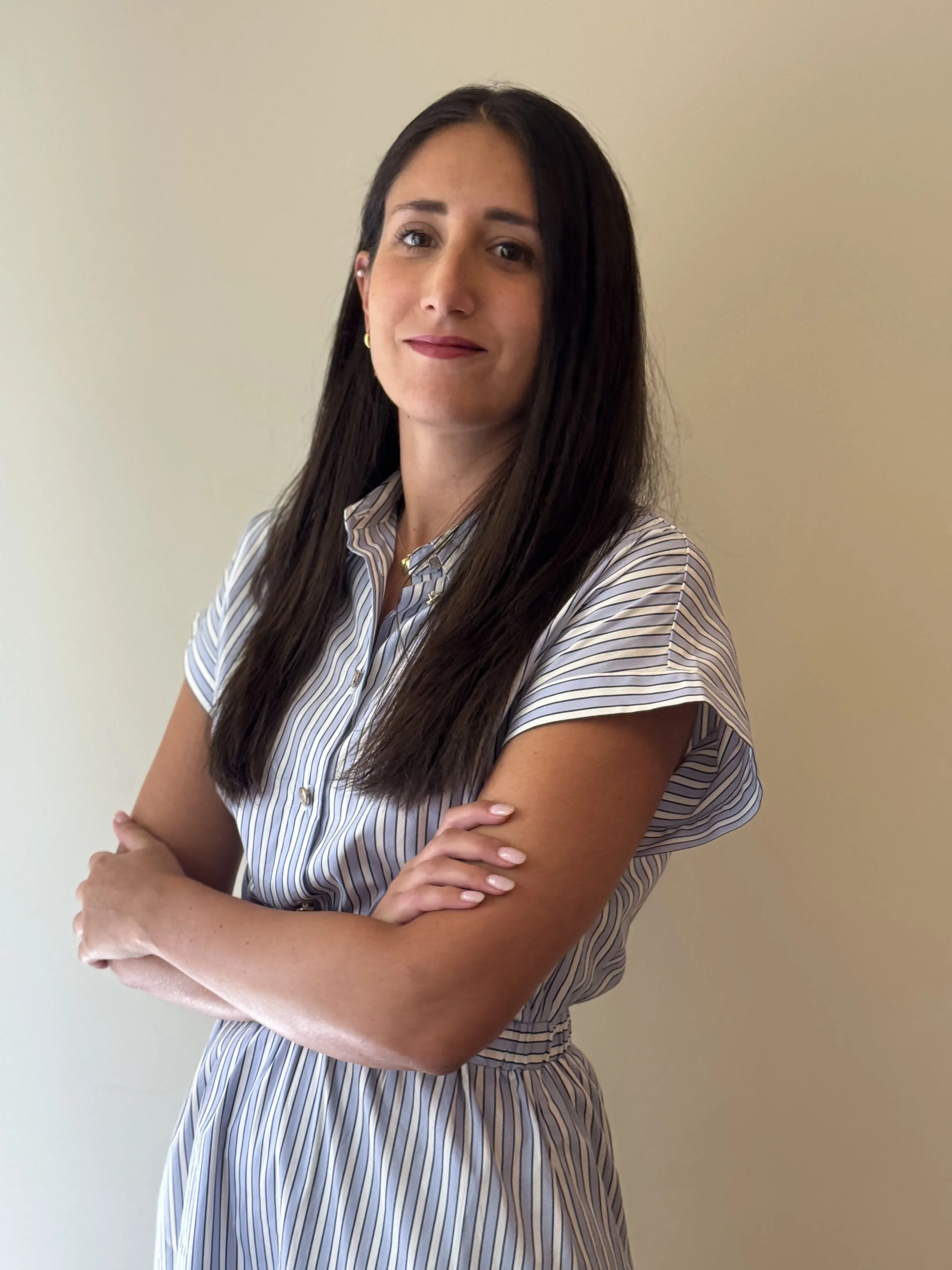 Foto della psicologa, Donna con capelli lunghi, sorriso, vestito a righe, mani incrociate, sfondo neutro.