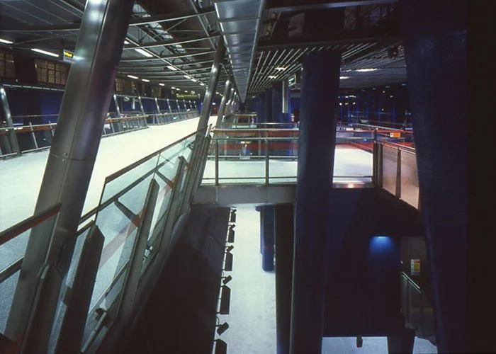 A view of the interior of North Greenwich underground station showing the slit levels from the ground floor with glass railings and metal beams looking down towards the underground train platform level.