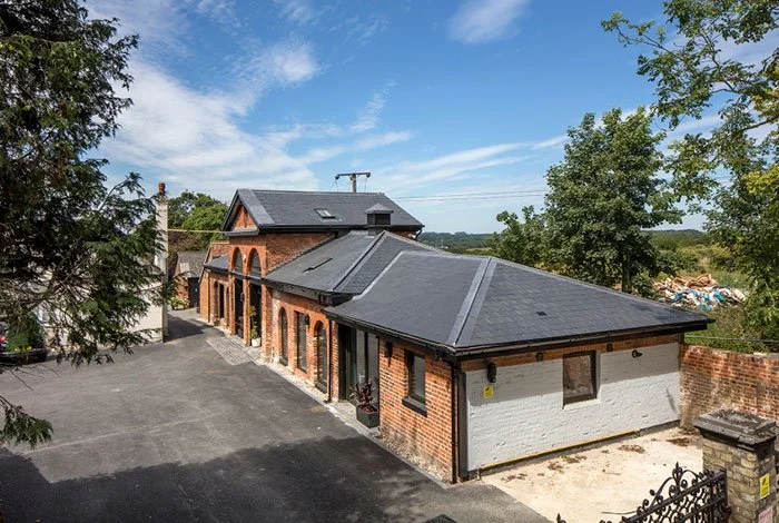 The Mascalls Stable Block brick building showing the dark grey slate roofs, large black framed windows, and surrounding greenery, viewed from above.