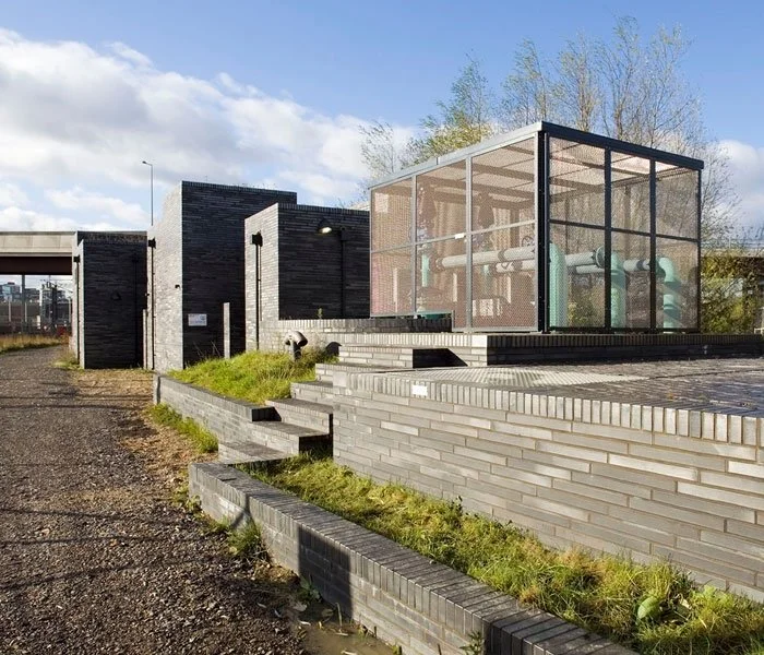 A side view of the Stratford Box Pumping Station in the main foreground with dark brick walls and a glass-enclosed structure housing pipes, surrounded by grassy areas and steps, under a partly cloudy sky.