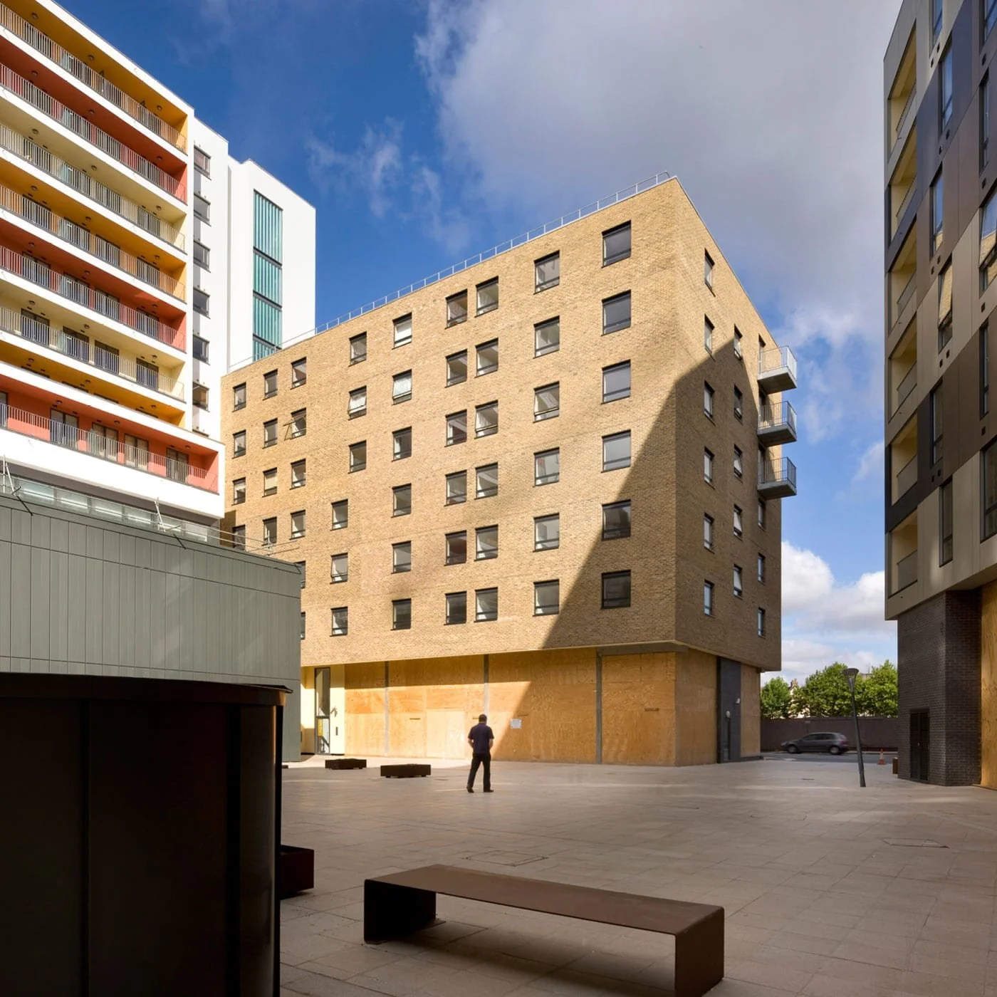 Cityscape with a modern beige brick building, surrounding colorful high-rises, and a man standing in an open plaza.