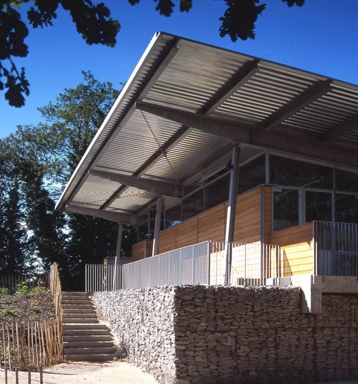 A side angle of Crystal Palace Park Renovation with metal roof, wooden siding, and glass walls, elevated on a stone foundation, with stairs leading up to a patio area.
