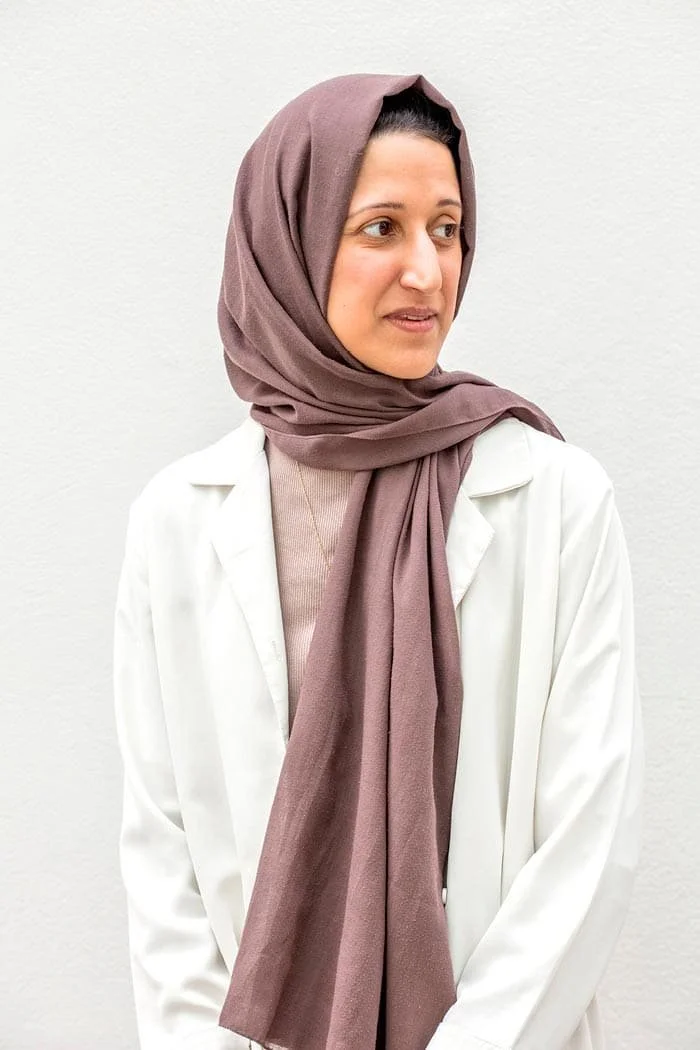 A woman wearing a light pink hijab and white lab coat, looking to the side, standing against a plain white wall.