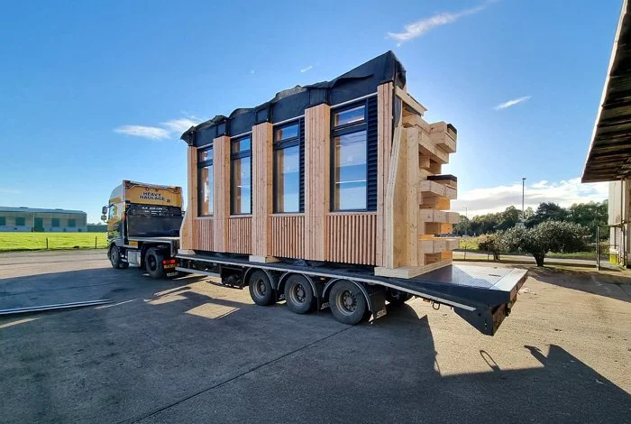 A small wooden house or building on a flatbed truck, being transported to a new location on a sunny day.