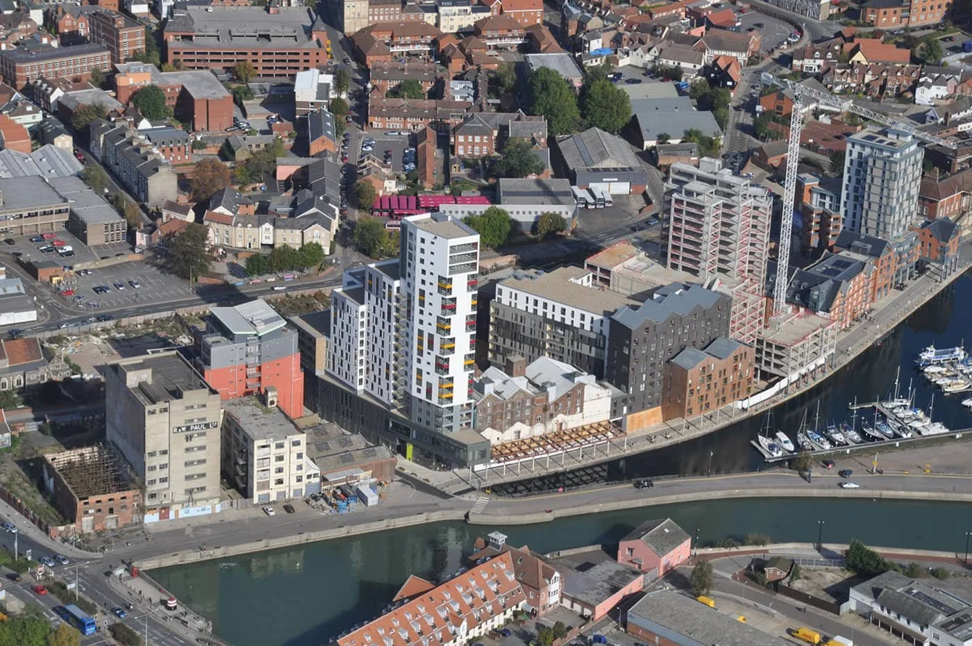 Aerial view of a city with modern high-rise buildings, a river with boats, and older smaller buildings and houses, with streets and parking lots.