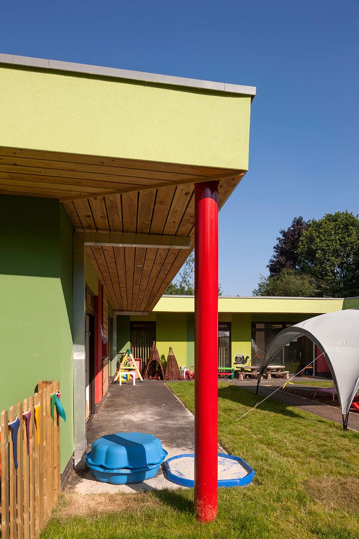 Enchanted Wood Nursery colourful outdoor area of a childcare or preschool with green walls and a red pole. There are toys, playground equipment, and a small splash pad on the grass. A white canopy provides shade, and a wooden fence.