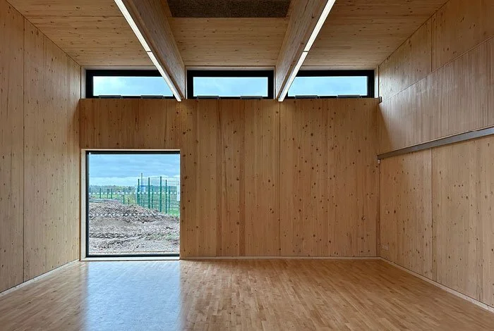Inside Education Sandpit building, an empty room with wooden walls and floor, large window and door showing an outdoor landscape, and wooden beams on ceiling.