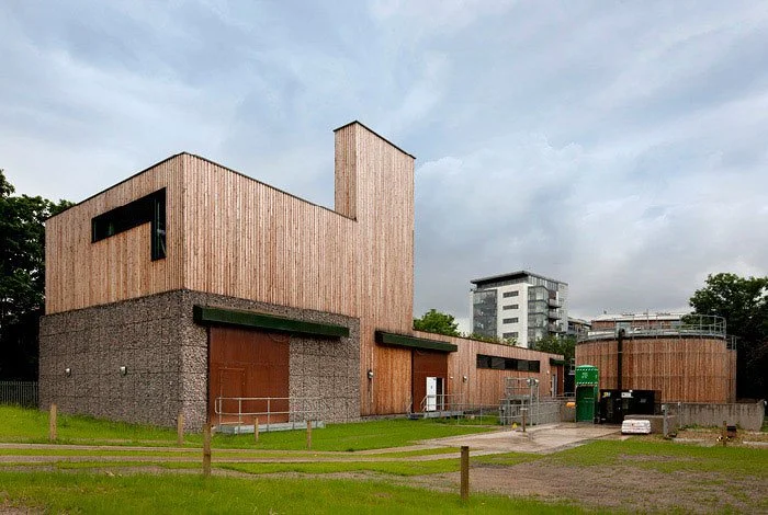 Modern building with wooden and concrete exterior, situated in a grassy area with trees and an urban background, under a cloudy sky.