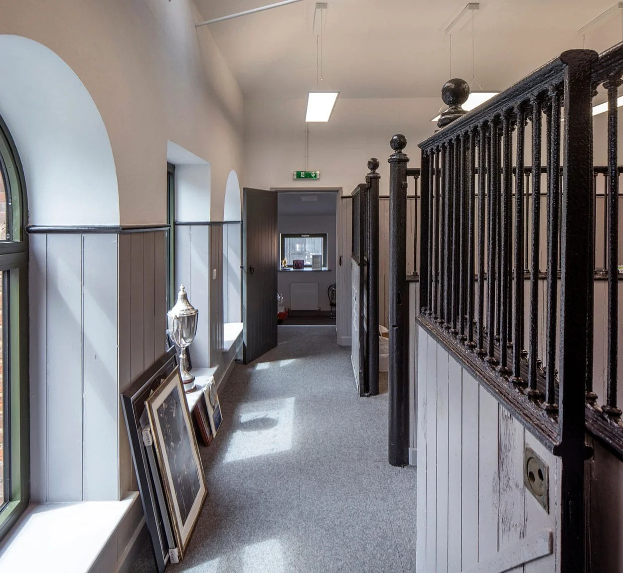 Hallway with arched windows, framed pictures leaning against wall, trophies, and a wooden door leading to another room with a window and chair.