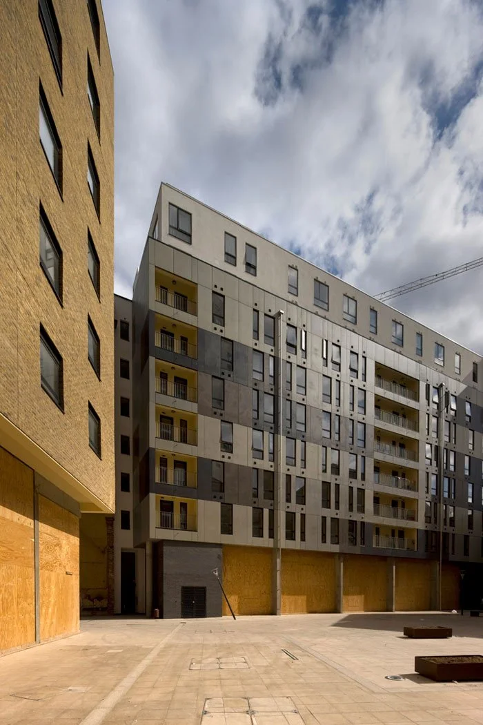 Modern multi-story residential building with balconies and a decorative facade, surrounded by an open paved area under a cloudy sky.