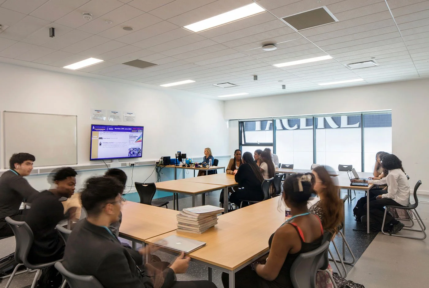 Students attending a class in a modern classroom, sitting at desks with laptops and notebooks, with a teacher at the front near a large screen displaying a presentation.