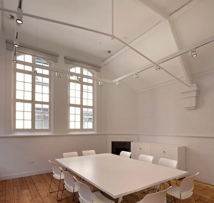 Empty conference room with white walls, large windows, and a white rectangular table surrounded by white chairs.
