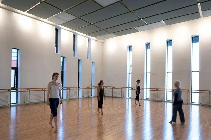 Four women practicing ballet in a spacious, well-lit dance studio with large windows and wooden floors.
