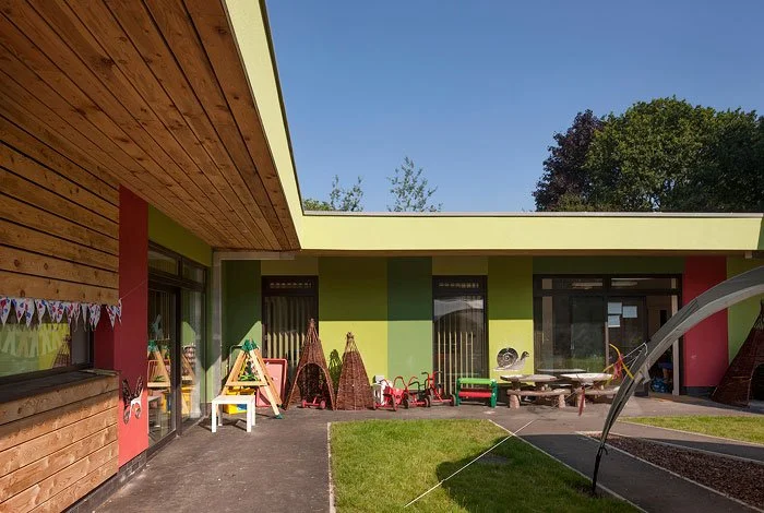Enchanted Wood Nursery outdoor area of a modern building with playground equipment, chairs, and a grassy space under a clear blue sky.