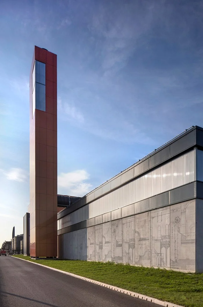 Modern industrial building with a tall, narrow tower and mural art on the lower wall, set against a blue sky.