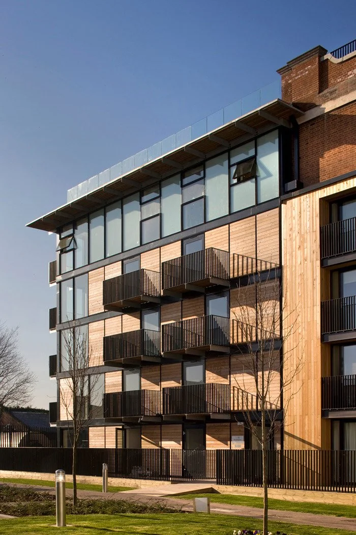 A view of the The Pump House extension with glass windows and black balconies, featuring wood paneling exterior, situated in a landscaped area with trees and a fence.