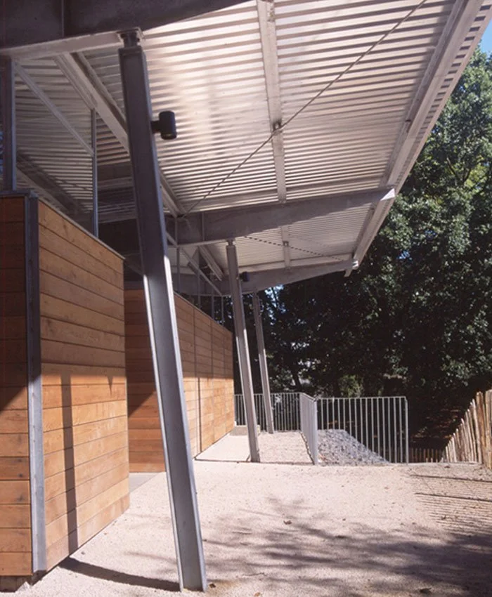A close-up view looking down the sire of Crystal Palace Park Renovation, a metal and glass roof overhang, supported by angled metal beams, and a wooden wall beneath. There is a gravel pathway bordered by a metal railing and trees in the background.