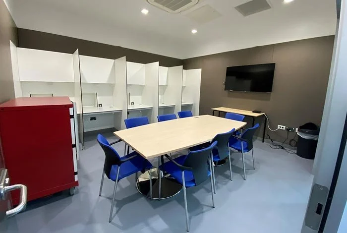A conference room with a rectangular table surrounded by eight blue chairs, white cubicle workstations in the background, a wall-mounted TV, a small table, and a red filing cabinet.