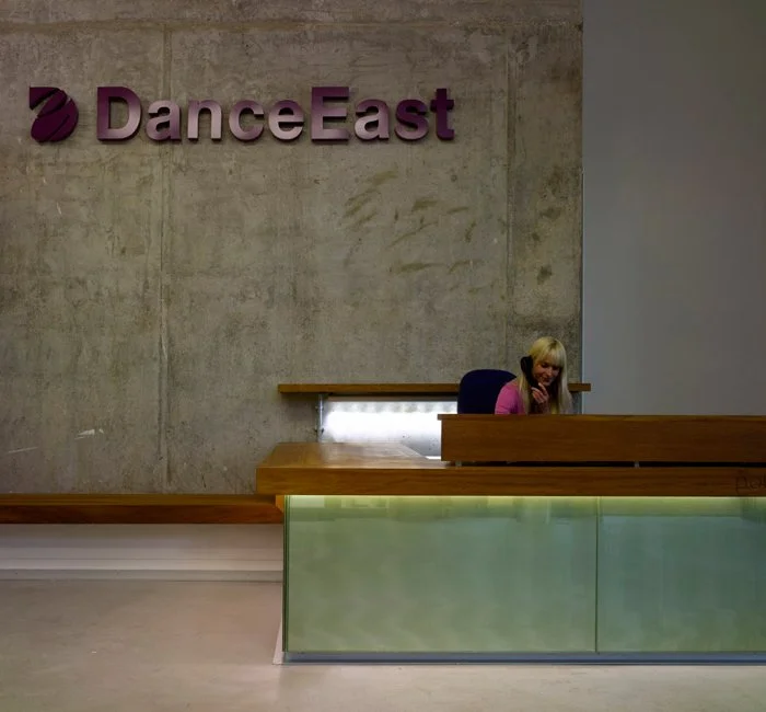 Reception area with a woman sitting at a desk, concrete wall background, 'DanceEast' sign on the wall, modern decor with wooden and frosted glass elements.