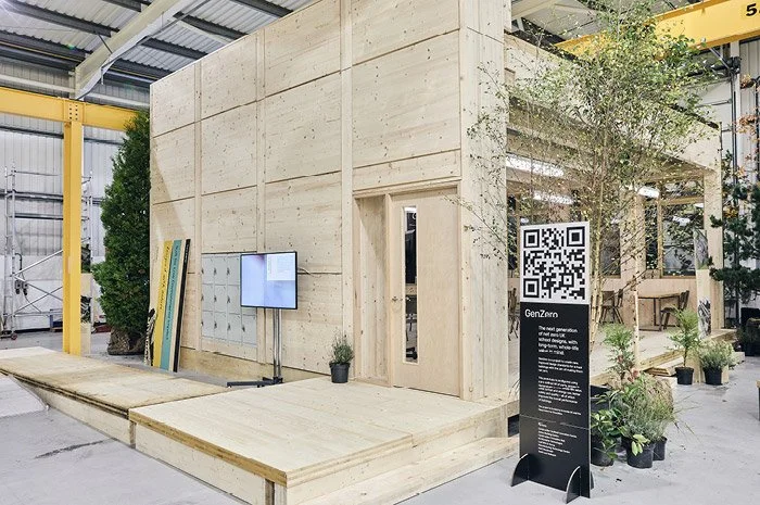A modern, wooden structure with a small outdoor stage or platform, surrounded by potted plants, with a QR code sign and a TV screen outside. The setting appears to be an industrial or exhibition space.