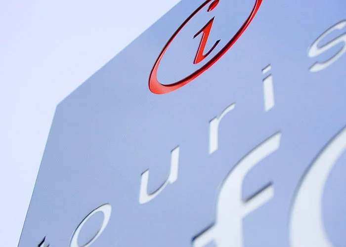 A close-up view of the Tourist Information Centre external main metal-cut lettering sign panel, with the distinctive italic 'i' in a circle in bright red, with the main remaining lettering in white in depth of field in the foreground.