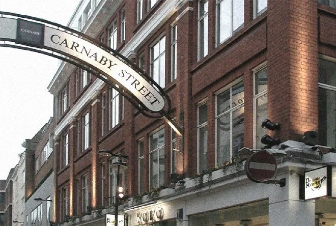 Street sign reading 'Carnaby Street' hanging above a brick building with multiple windows, a streetlamp, and a 'no entry' traffic sign in an urban setting.