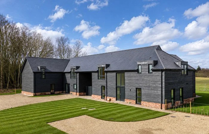Modern black house with large windows and a sloped roof, surrounded by a neatly maintained lawn and gravel pathway, under a partly cloudy sky.