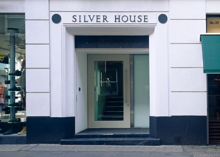 The main entrance of a building with the signage for Silver House in traditional style on the stone wall facade, with glass doors and a staircase inside, surrounded by white walls.