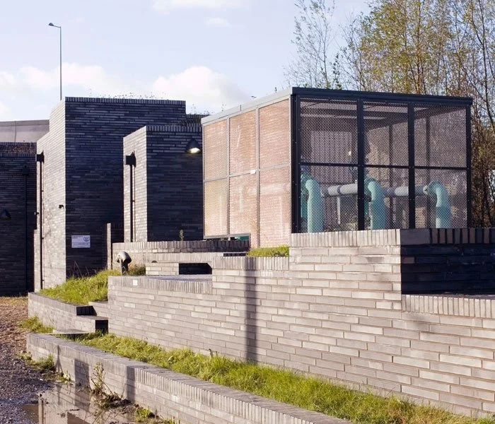 A close up of the Stratford Box Pumping Station's modern building with brick and concrete walls, featuring a glass-enclosed structure with several blue and white pipes inside, surrounded by grass and steps.