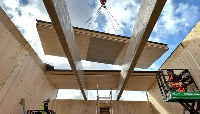 Construction workers installing a large wooden panel between two wooden walls, while a crane lifts it overhead on a construction site.