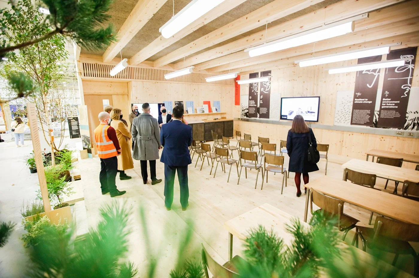People attending a presentation or exhibit in a modern indoor space with wooden walls and ceiling, green plants, and seating arranged in rows facing the front where a monitor displays information.